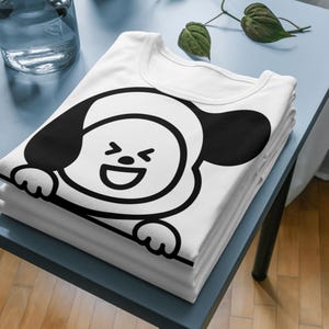 Puede incluir: Una pila de camisetas blancas con un dise&ntilde;o de personaje de dibujos animados en blanco y negro. El dise&ntilde;o es una cara sonriente con grandes orejas y patas negras. Las camisetas est&aacute;n cuidadosamente dobladas sobre una superficie azul.