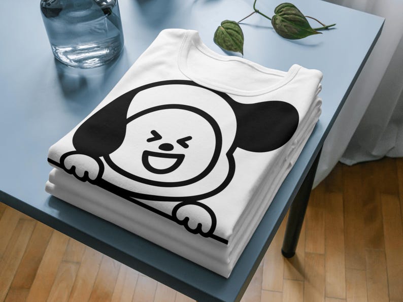 Puede incluir: Una pila de camisetas blancas con un dise&ntilde;o de personaje de dibujos animados en blanco y negro. El dise&ntilde;o es una cara sonriente con grandes orejas y patas negras. Las camisetas est&aacute;n cuidadosamente dobladas sobre una superficie azul.
