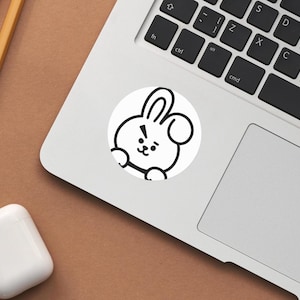Puede incluir: Una pegatina blanca con un personaje de conejo de dibujos animados con contornos negros, colocada en un port&aacute;til. El conejo tiene una expresi&oacute;n de sorpresa. El port&aacute;til es plateado y descansa sobre una superficie marr&oacute;n. Tambi&eacute;n se ven palillos de madera y una funda blanca para auriculares.