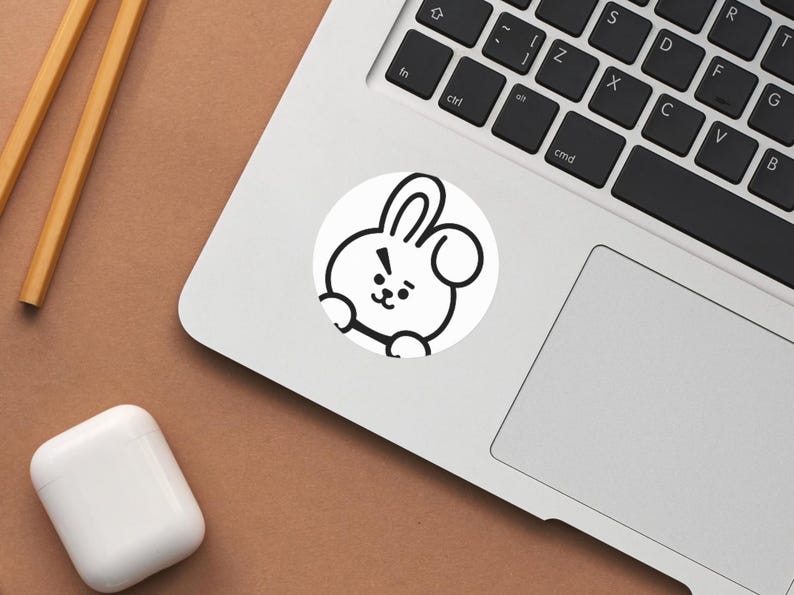 Puede incluir: Una pegatina blanca con un personaje de conejo de dibujos animados con contornos negros, colocada en un port&aacute;til. El conejo tiene una expresi&oacute;n de sorpresa. El port&aacute;til es plateado y descansa sobre una superficie marr&oacute;n. Tambi&eacute;n se ven palillos de madera y una funda blanca para auriculares.