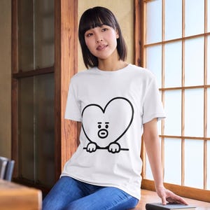 Puede incluir: Camiseta blanca con un personaje negro en forma de coraz&oacute;n con cara y manos. El personaje est&aacute; delineado en negro. La camiseta se usa con jeans azules.