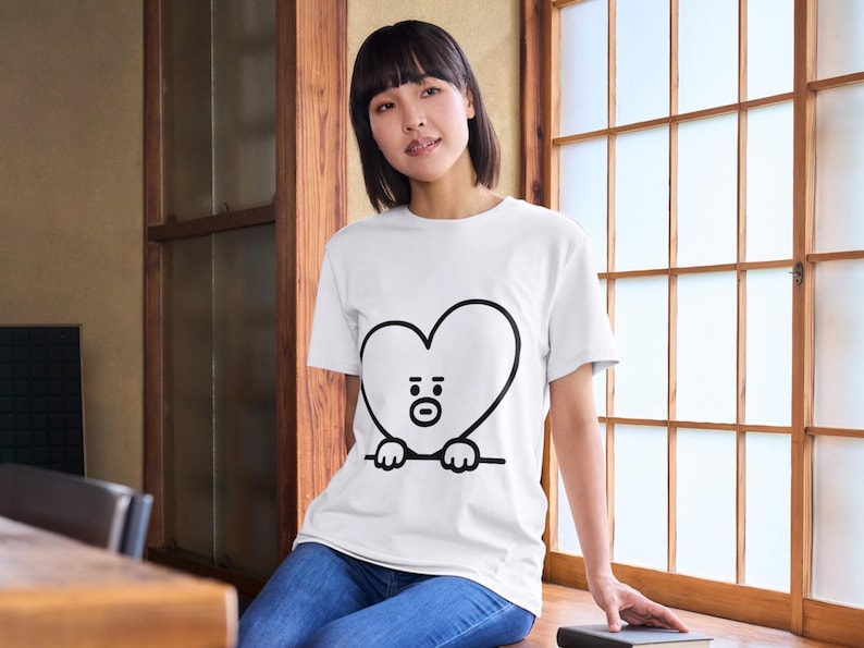 Puede incluir: Camiseta blanca con un personaje negro en forma de coraz&oacute;n con cara y manos. El personaje est&aacute; delineado en negro. La camiseta se usa con jeans azules.