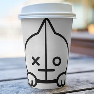 Puede incluir: Una taza de caf&eacute; de papel blanca con tapa blanca. La taza presenta un dise&ntilde;o de personaje de dibujos animados negro con una X para un ojo y un ojo circular en el otro lado. La taza est&aacute; sobre una superficie de madera.