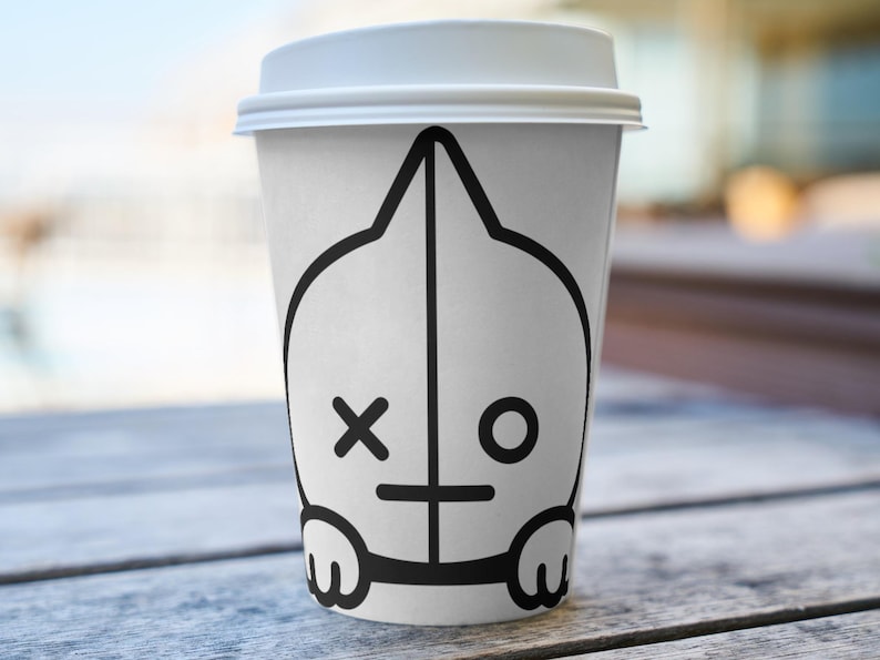 Puede incluir: Una taza de caf&eacute; de papel blanca con tapa blanca. La taza presenta un dise&ntilde;o de personaje de dibujos animados negro con una X para un ojo y un ojo circular en el otro lado. La taza est&aacute; sobre una superficie de madera.