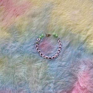 May include: A white beaded bracelet with the words "Agent Whiskey" spelled out in black letters. The beads are separated by green beads with a heart shape.