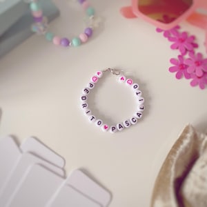 May include: A white beaded bracelet spelling "PEDRITO PASCAL" in black letters, with pink heart beads. The image also shows pink sunglasses, a beaded bracelet, and pink flower decorations, all on a white surface.