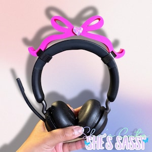 May include: Black headset with a pink bow and a heart-shaped gem. The headset has a microphone and is held by a hand. The text "She's Sassy" is visible in the bottom right corner.