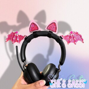 May include: Black headphones with pink bat wings and cat ears. The wings and ears are embellished with glitter and rhinestones. The text "She's Sassy" is visible in the bottom right corner.