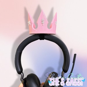 Può includere: Un supporto per cuffie a forma di corona rosa con dettagli argentati è montato su una cuffia nera. La corona ha un design a tiara con una chiave e dettagli floreali. La cuffia è nera e ha un microfono.