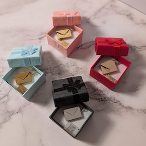 May include: A selection of small gift boxes in shades of pink, red, blue, and black, each tied with a matching ribbon. Inside each box is a necklace with a small envelope pendant. The boxes are open, displaying the jewellery.