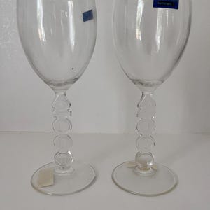 Könnte beinhalten: Zwei klare Weingläser mit einem einzigartigen Stiel-Design. Die Stiele weisen eine Reihe von verbundenen Kreisen auf. Jedes Glas hat einen runden Fuß und ein Etikett mit der Aufschrift "MILLENNIUM Luminarc".
