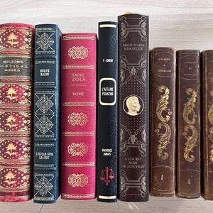 Può includere: Una collezione di otto libri con copertina rigida disposti in verticale, con dorsi vari in rosso, blu scuro, nero e marrone con scritte dorate e motivi decorativi, inclusi titoli come Milton's Poetical Works e L'Affaire Pranzini.