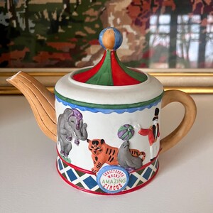 Peut inclure: Théière en céramique décorative sur le thème du cirque. Elle présente un éléphant, un tigre, un phoque et un maître de cérémonie. Le couvercle est rayé rouge, vert et blanc, et l'anse et le bec sont beiges. La base porte l'inscription "Amazing Circus".