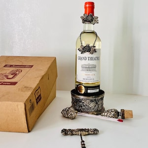 Puede incluir: Una botella de vino Grand Theatre Burdeos con un collar y una base decorativos con temática de uvas. También hay un sacacorchos y un tapón de botella de color plateado. La botella tiene una tapa roja y se exhibe con una caja de cartón.
