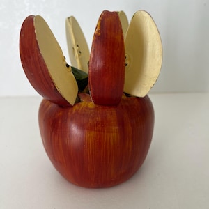 May include: A decorative apple-shaped object featuring a red and yellow painted base with several upright, curved, cream-coloured slices protruding from the top. The slices have a smooth finish and are arranged to resemble a cut apple.