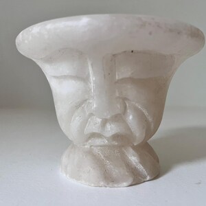 Könnte beinhalten: Eine geschnitzte, cremefarbene Skulptur eines Gesichts mit geschlossenen Augen und Bart. Die Skulptur hat eine ausgestellte Oberseite und einen abgerundeten Sockel. Das Gesicht ist detailliert mit geschnitzten Merkmalen, und das Gesamtdesign ist glatt und organisch.