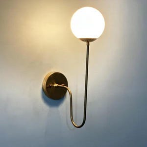 May include: A modern wall sconce featuring a spherical, frosted glass shade. The light fixture has a brass finish and a curved arm that connects to a circular wall mount. The design is minimalist and elegant, suitable for various interior styles.