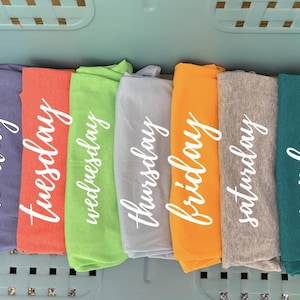 May include: Seven folded t-shirts in a row, each a different color: purple, coral, green, gray, orange, gray, and teal. Each shirt has a day of the week written in white script: Monday, Tuesday, Wednesday, Thursday, Friday, Saturday, and Sunday.