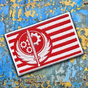 May include: A rectangular embroidered patch featuring a red and white striped background. Centered on the patch is a white circular emblem depicting a vertical sword flanked by wings, with three interconnected gears positioned above the sword hilt.