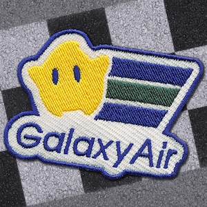 May include: Embroidered patch featuring a yellow star with blue eyes and a blue and green striped design. The patch has the text "GalaxyAir" in blue, outlined in white. The patch is on a black and white checkered background.
