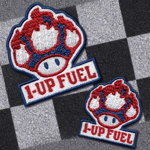Pode incluir: Dois patches bordados com um design de cogumelo vermelho e branco com detalhes em azul. Cada patch tem uma faixa retangular branca com o texto "1-UP FUEL" em vermelho. Os patches estão em um fundo quadriculado preto e cinza.
