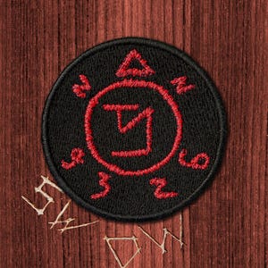 May include: A circular black embroidered patch featuring a red stitched border and a central red symbol enclosed within a circle. The design includes red characters arranged around the perimeter, with a distinct red geometric shape in the center.