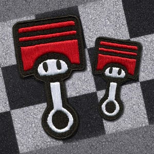 May include: Two embroidered patches in the shape of a piston, one large and one small. The pistons are black with red tops and white accents. The background is a black and white checkered pattern.