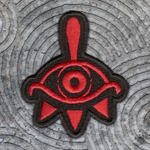May include: An embroidered patch featuring a stylized eye design with a central pupil, a curved eyelid, and three downward-pointing triangular shapes at the base. The design is rendered in vibrant red thread against a contrasting black background.