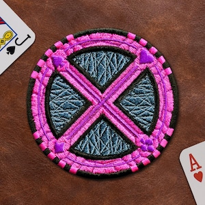May include: A circular embroidered patch featuring a dark blue textured center divided by a bright pink X-shaped cross. The border is composed of alternating black and pink segments, accented with small purple suit symbols at the cardinal points.