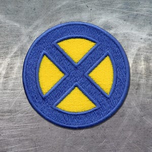 May include: A circular embroidered patch featuring a blue border and a blue cross shape that divides the center into four yellow triangular sections. The stitching is dense and uniform, creating a textured surface with clear, defined edges.