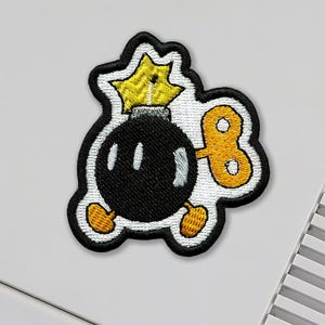 May include: Embroidered patch featuring a cartoon bomb character with a black body, yellow star fuse, and wind-up key. The patch has a white border with black outlining and yellow feet.