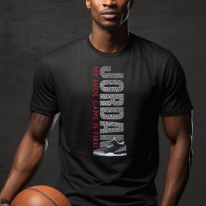 Puede incluir: Una camiseta negra con un gr&aacute;fico blanco de la palabra "JORDAN" formado por patrones de suela de zapato. El texto "MY SHOE GAME IS FIRE!" est&aacute; impreso en rojo en el lateral del gr&aacute;fico. Un zapato de baloncesto blanco y negro est&aacute; representado debajo del gr&aacute;fico.