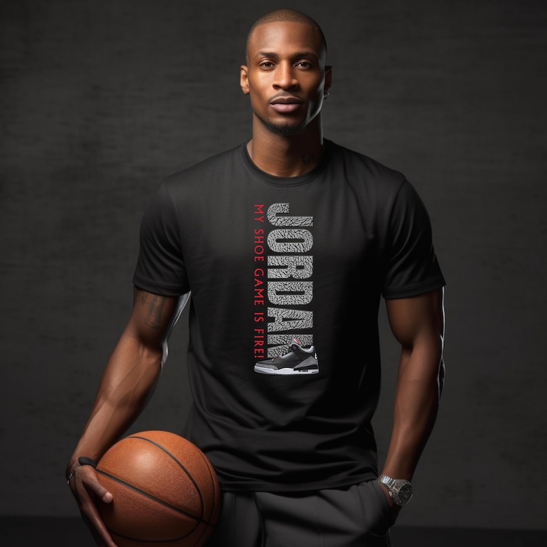 Puede incluir: Una camiseta negra con un gr&aacute;fico blanco de la palabra "JORDAN" formado por patrones de suela de zapato. El texto "MY SHOE GAME IS FIRE!" est&aacute; impreso en rojo en el lateral del gr&aacute;fico. Un zapato de baloncesto blanco y negro est&aacute; representado debajo del gr&aacute;fico.