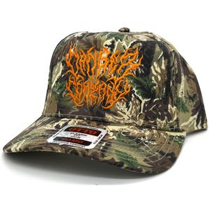 Op de afbeelding: Een camouflage baseball cap met een oranje, gestileerd tekstontwerp. De pet heeft een gebogen klep en een label met de tekst "OTTO" en "CLASSIC FIT CAMO". De hoed is gemaakt van een premium stof.