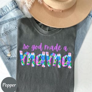 May include: A gray t-shirt with the text "so god made a mama" in purple letters with a floral design. The word "mama" is in large letters and has a floral pattern.