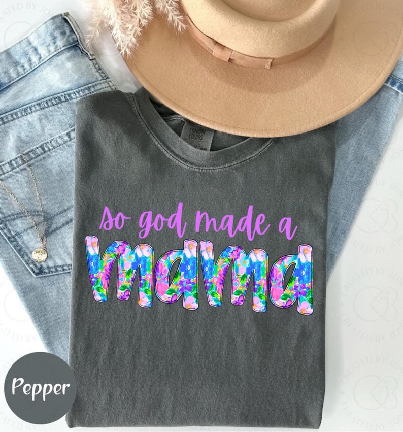 May include: A gray t-shirt with the text "so god made a mama" in purple letters with a floral design. The word "mama" is in large letters and has a floral pattern.