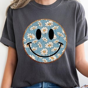 Puede incluir: Camiseta gris oscuro con un dise&ntilde;o de cara sonriente grande. La cara sonriente tiene un fondo azul con flores de margarita blancas, ojos negros y una sonrisa negra. El borde de la cara sonriente es dorado.