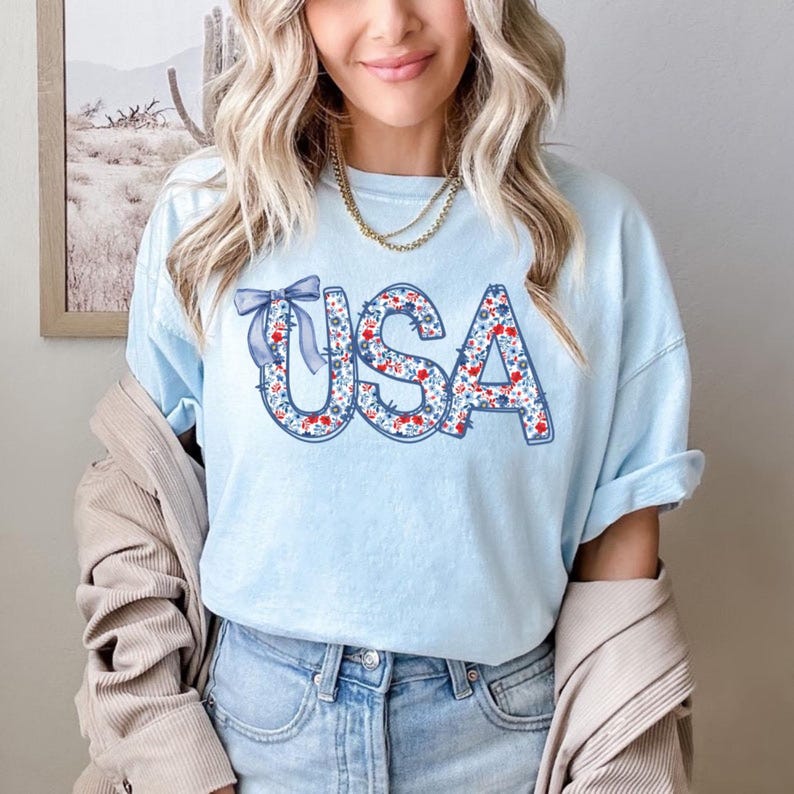 以下が含まれることがあります： 薄い青色のTシャツに、赤、白、青の色の花柄で「USA」と綴られています。青いリボンが単語の上にあります。