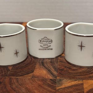 May include: Three white ceramic napkin rings with a silver trim and starburst designs. One ring features the text "Fukagawa Arta Handpainted Made in Japan". The napkin rings are cylindrical and sit on a wooden surface.