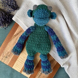 May include: A crocheted plush toy in the shape of a monster with green and blue fur. The toy has large, round eyes and a small, pointed nose. It has long, thin arms and legs.