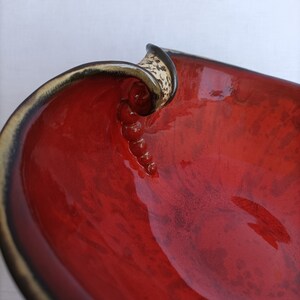 A heart-shaped ceramic bowl with a shiny red glaze on the inside and a brown rim. The exterior is stone-like in brown and beige. The curve of the heart is like a spiral and several red balls descend in a row below it.