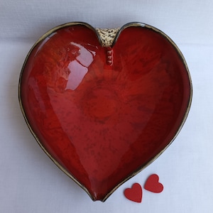 A heart-shaped ceramic bowl with a shiny red glaze on the inside and a brown rim. The exterior is stone-like in brown and beige. The curve of the heart is like a spiral and several red balls descend in a row below it.