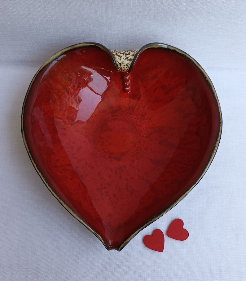 A heart-shaped ceramic bowl with a shiny red glaze on the inside and a brown rim. The exterior is stone-like in brown and beige. The curve of the heart is like a spiral and several red balls descend in a row below it.