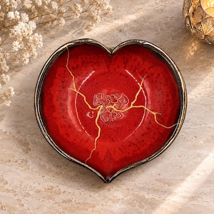 May include: A heart-shaped ceramic dish with a vibrant red glaze and a dark, metallic-toned rim. The center features a gold-colored crack repair pattern resembling kintsugi, with small embossed heart shapes clustered near the center point.