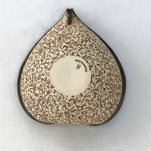 A heart-shaped ceramic bowl with a shiny red glaze on the inside and a brown rim. The exterior is stone-like in brown and beige. The curve of the heart is like a spiral and several red balls descend in a row below it.
