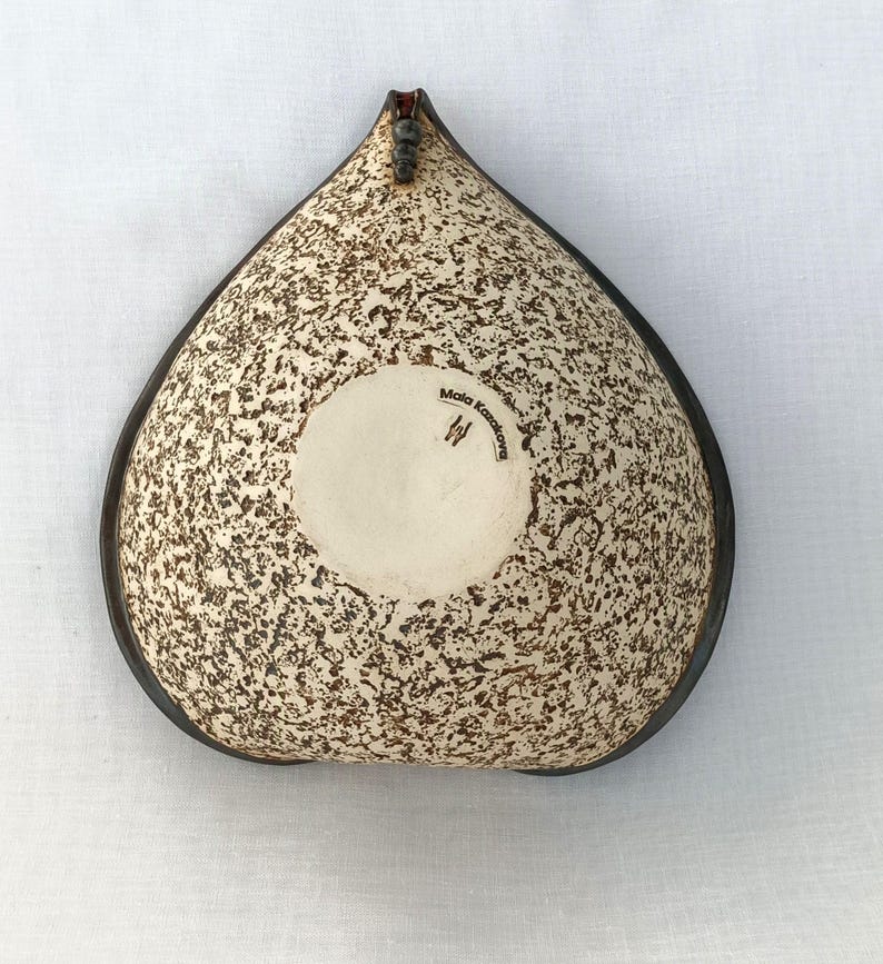 A heart-shaped ceramic bowl with a shiny red glaze on the inside and a brown rim. The exterior is stone-like in brown and beige. The curve of the heart is like a spiral and several red balls descend in a row below it.