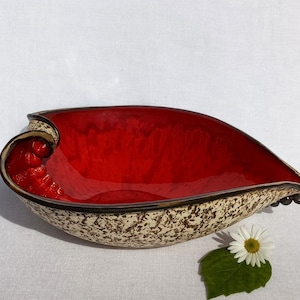 A ceramic bowl in the shape of a heart with a shiny red glaze on the inside and a brown rim. The exterior is stone-like in brown and beige. The curve of the heart is like a spiral and several red balls descend in a row below it. The sharp tip of the heart also ends with a spiral and several balls in a brown-copper color.