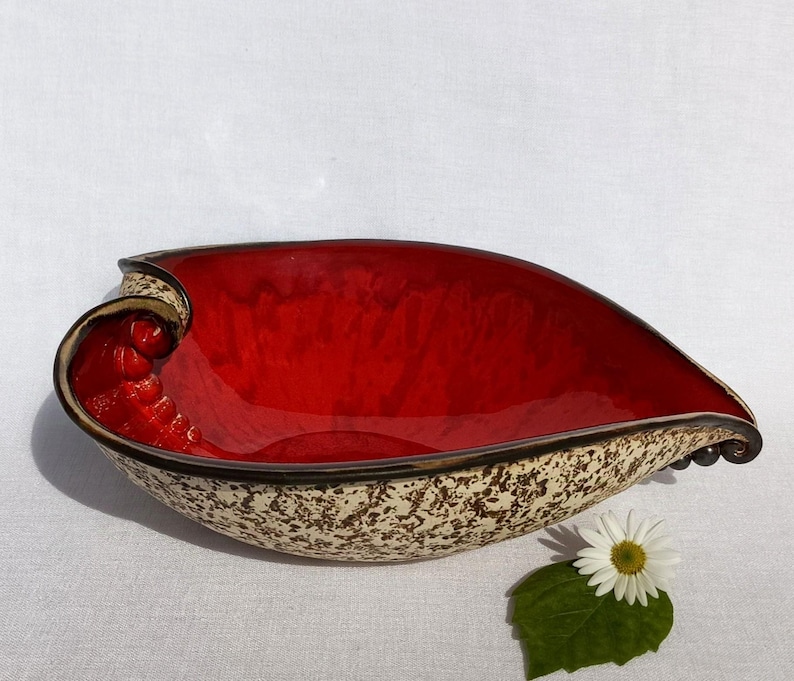 A ceramic bowl in the shape of a heart with a shiny red glaze on the inside and a brown rim. The exterior is stone-like in brown and beige. The curve of the heart is like a spiral and several red balls descend in a row below it. The sharp tip of the heart also ends with a spiral and several balls in a brown-copper color.