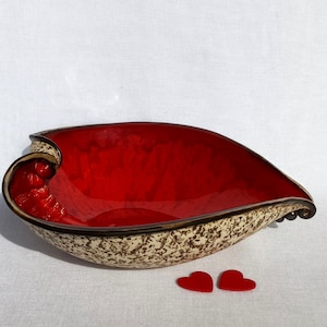 A heart-shaped ceramic bowl with a shiny red glaze on the inside and a brown rim. The exterior is stone-like in brown and beige. The curve of the heart is like a spiral and several red balls descend in a row below it.