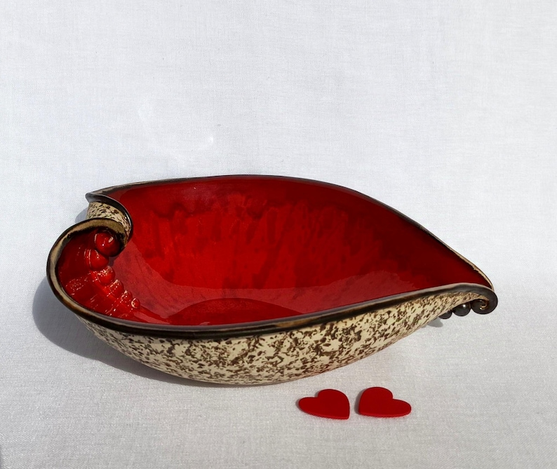 A heart-shaped ceramic bowl with a shiny red glaze on the inside and a brown rim. The exterior is stone-like in brown and beige. The curve of the heart is like a spiral and several red balls descend in a row below it.